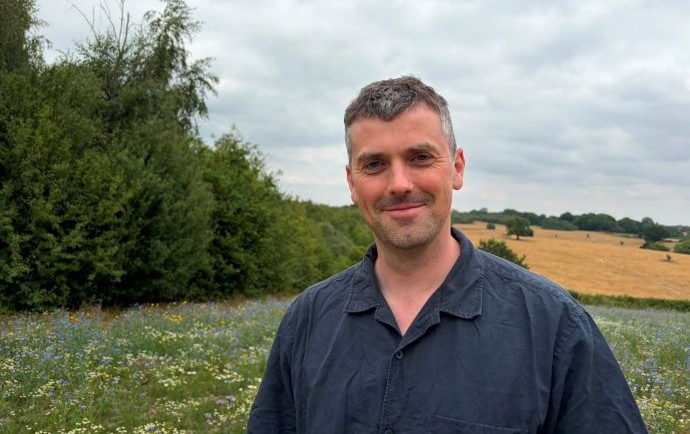 Jack McCrickard stands in a meadow with trees behind him, smiling at the camera