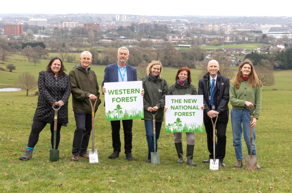 A group of Western Forest staff and partners stand on a hill at Ashton Court, holding spades and smiling. They have a sign that reads 'Western Forest - The new national forest'
