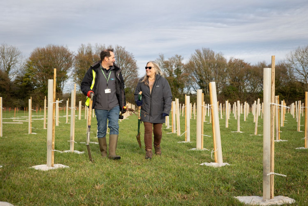 Two people walking towards the camera, talking to each other and smiling. They are in a field, surrounded by stakes and guarded saplings