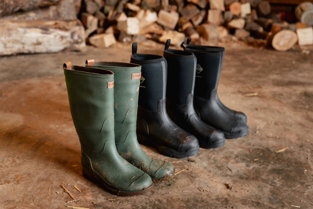 Three pairs of wellington boots lined up on the ground next to a pile of chopped logs