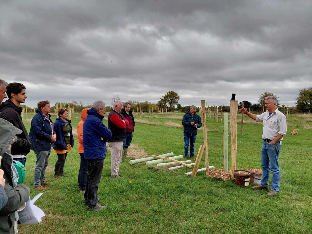 A man stands next to a sapling tree that is being planted in a field, a crowd of people listening.